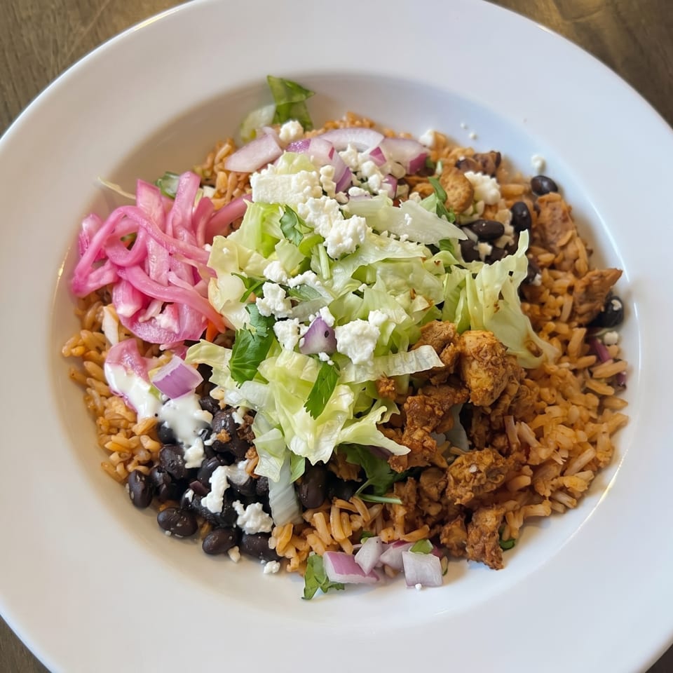 A colorful rice bowl with chicken, black beans, lettuce, pickled onions, and feta cheese.